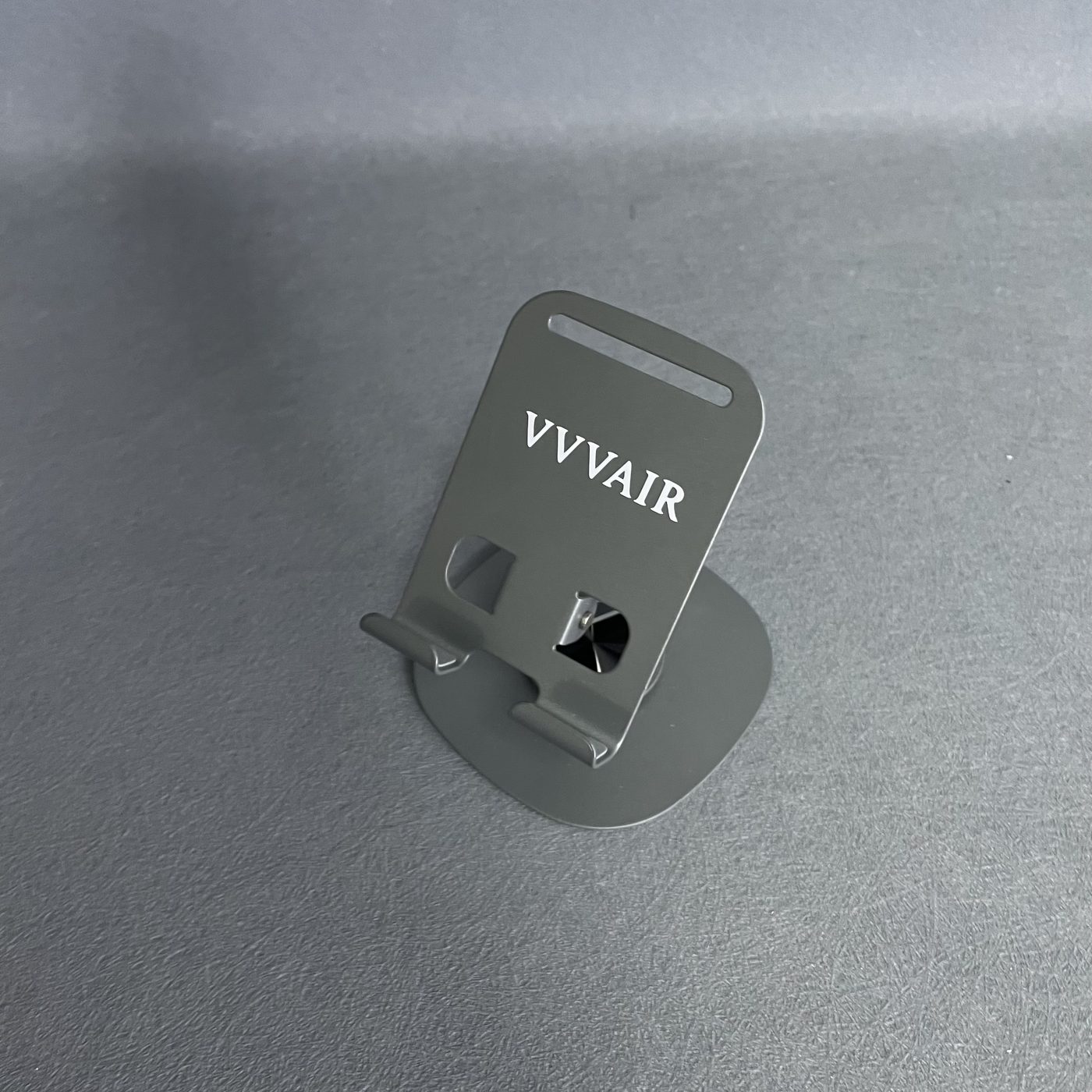 The Space Gray mobile phone stand standing upright on a dark grey textured surface, with the VVVAIR mark printed in white across the support plate